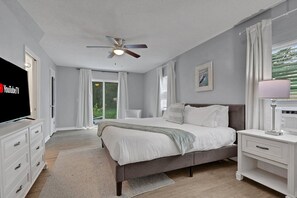 A bedroom with a bed, a ceiling fan, a TV, a bedside table with a lamp, and a window with curtains.