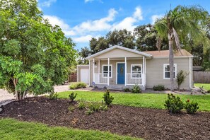 A single-story house with a front porch, a blue door, and a small garden area with mulch and plants. - Home w/ Fenced Yard, Hot tub, Grill & 3 King Beds! (Sarasota)