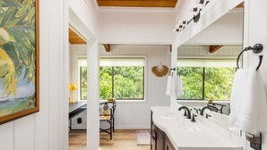 A bathroom with white walls, wooden ceiling, and a large mirror. There are two sinks, a window with a view of greenery, and a chair with a yellow lamp.