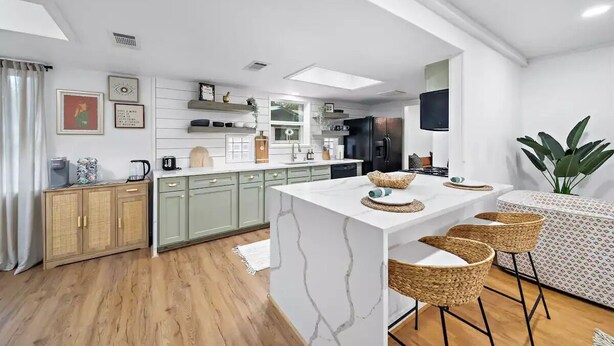 Una cuina moderna amb una illa de marbre, armaris de color verd clar i una zona de menjador amb cadires de vimet.