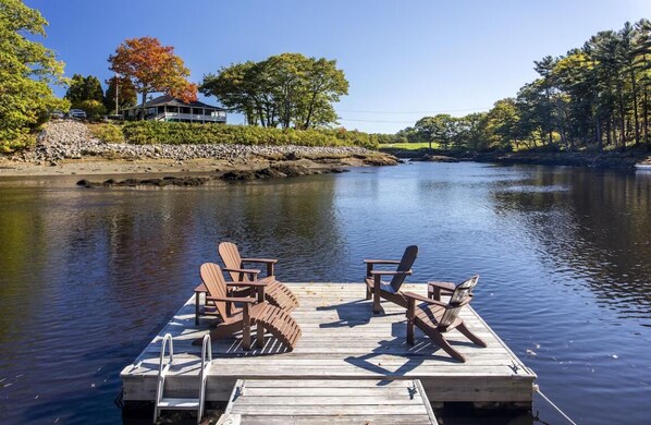 Une jetée en bois avec deux chaises Adirondack, donnant sur une rivière paisible, entourée d’arbres et avec une maison en arrière-plan.