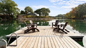 Un muelle de madera con dos sillas Adirondack y una mesa, con vista a un lago tranquilo con árboles y una casa al fondo.