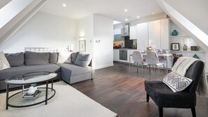 A modern living room with a sectional sofa, a glass coffee table, and a black armchair. The room features dark wooden flooring and white walls. - Farringdon/St Pancras penthouse (London)