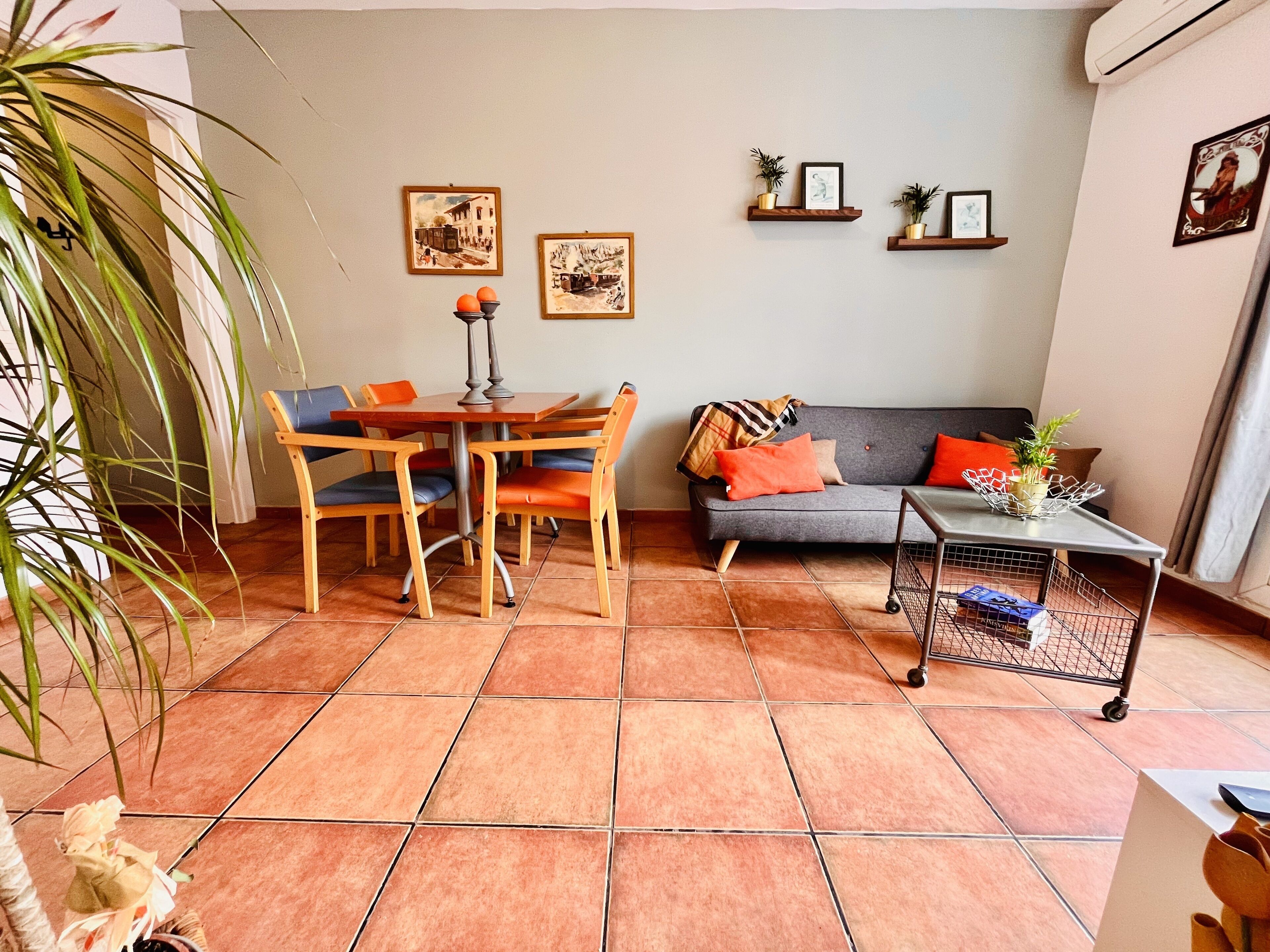 Un salon comprenant une table à manger, des chaises, un canapé et une table basse. Des tableaux encadrés ornent le mur, et une plante en pot est posée sur le sol.