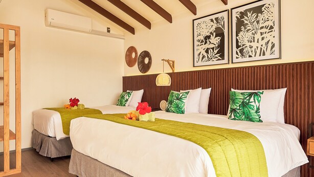 A hotel room with two beds, each with white bedding and green throw pillows. There are wall-mounted air conditioning units and framed botanical artwork above the beds.
