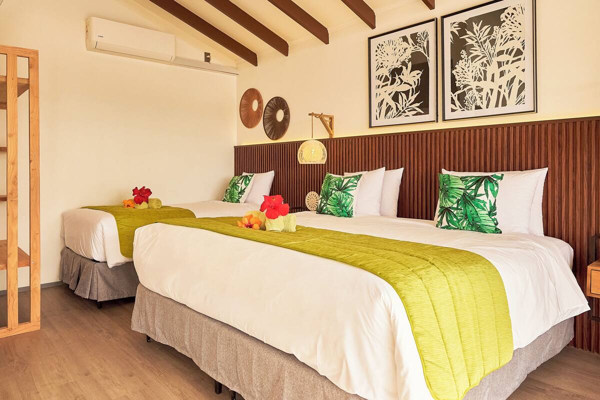Camera d'albergo con due letti, entrambi con biancheria da letto bianca e cuscini decorativi verdi. Sono presenti unità di condizionamento fissate al muro e stampe botaniche incorniciate sopra i letti.