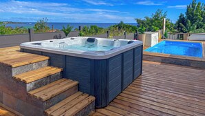 A hot tub and a pool on a wooden deck with a view of the ocean.
