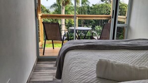 Un dormitorio con una cama grande, suelo de madera, balcón con mesa y sillas, y vistas a la vegetación.