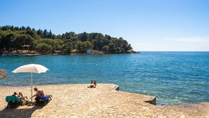 A stone-paved waterfront area with a white umbrella, two people relaxing, and a view of the sea and a forested island.
