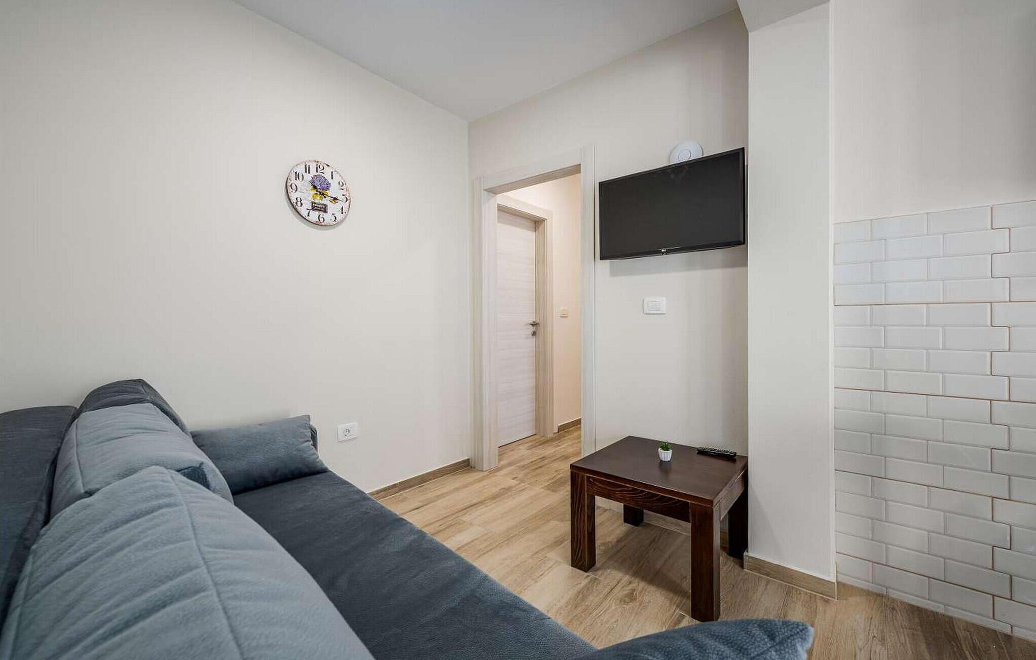 A living room with a grey sofa, a small wooden table, a wall-mounted TV, and a door leading to another room.