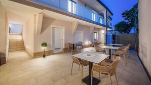 A spacious outdoor patio with multiple tables and wicker chairs, a staircase leading to an upper level, and a well-lit entrance.