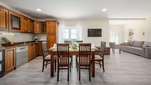 A modern kitchen with wooden cabinets, a dining table set for four, and a living area with a sofa and TV.