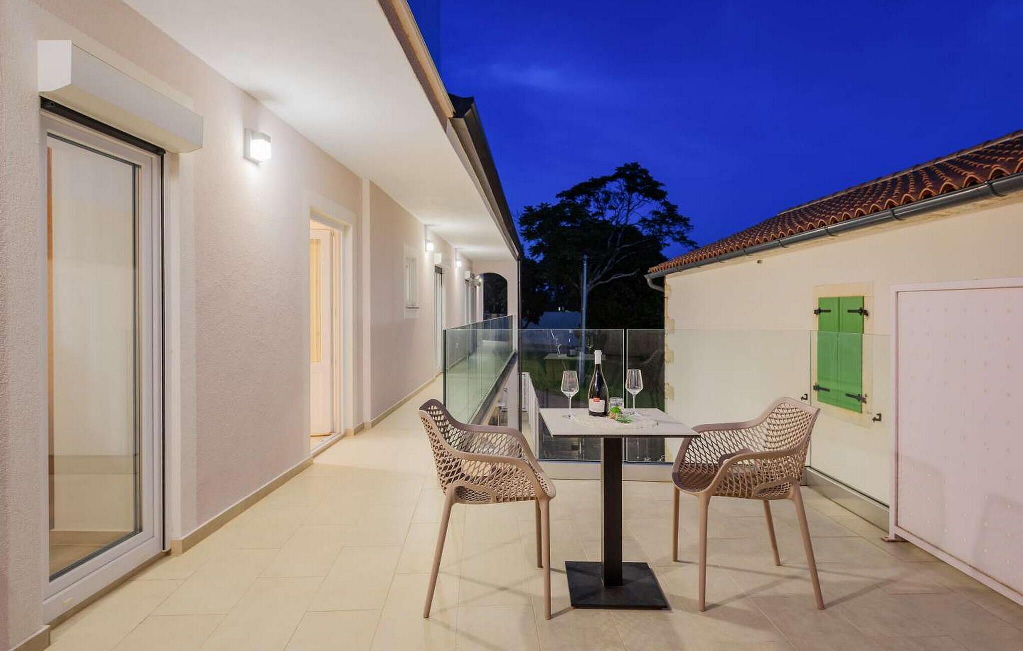 An outdoor patio with a glass table and two wicker chairs, a bottle of wine, and a green shuttered door.