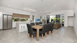 A modern kitchen with a central island, a dining area with a wooden table and chairs, and a living area with a sofa and large windows. - Seaside heated pool (Palm Cove)