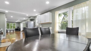 Un comedor con una mesa redonda y cuatro sillas, una cocina con armarios blancos y ventanas amplias con cortinas.