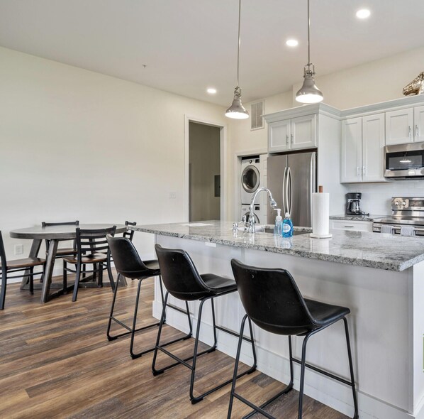 A modern kitchen with a central island, bar stools, a dining table, and a washing machine in the background.