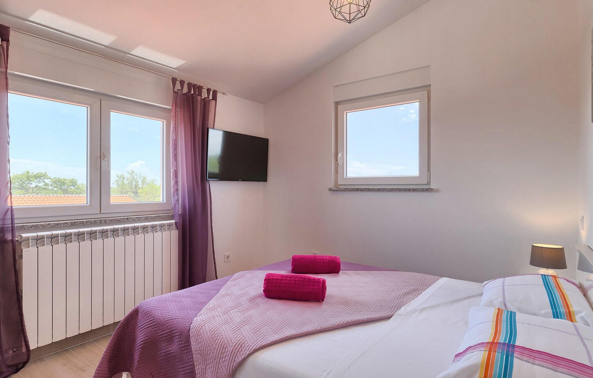 A bedroom with a bed, two pink towels, a TV on the wall, and a window with curtains.