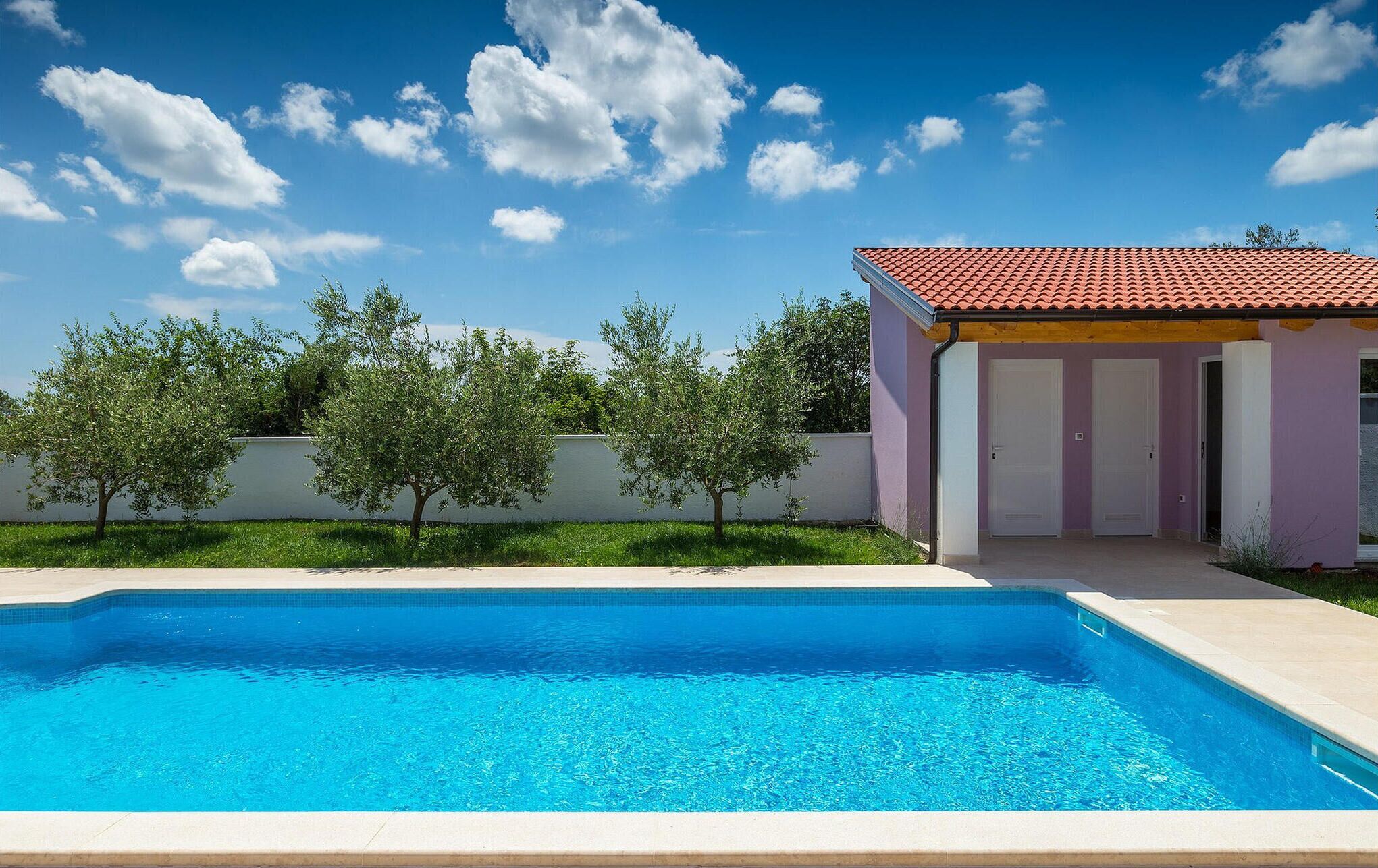A swimming pool with a clear blue water surface, surrounded by a low wall and a building with a red-tiled roof.