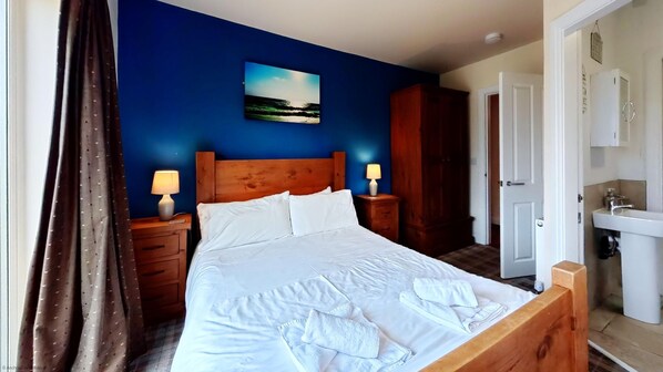 Un lit bien fait, avec du linge de lit et des serviettes blanches, une tête de lit en bois, deux lampes de chevet, un tableau représentant une scène de plage, une armoire et une salle de bain avec un lavabo et un miroir.