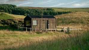 A rustic wooden cabin with a metal roof, surrounded by a wooden fence, set in a grassy field with a forested hill in the background. - Eco Off-Grid Luxury Cabin, Panoramic Views & Fire Pit In North Wales (Llangollen)