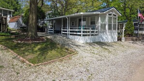 A small, single-story cabin with a porch and a covered outdoor area, surrounded by trees and a gravel driveway.