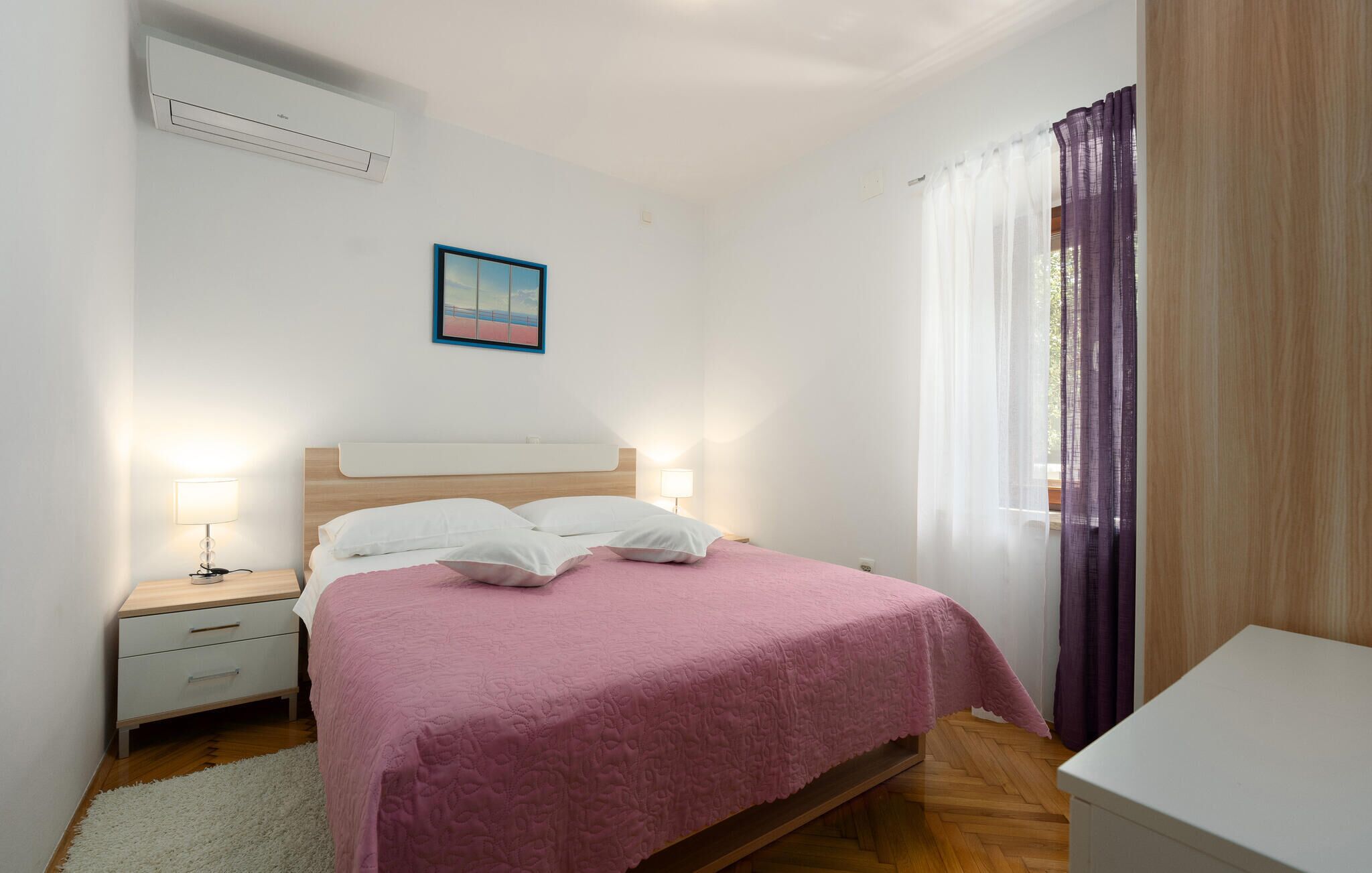 A bedroom with a bed, bedside tables, a nightstand, a wall-mounted air conditioner, a framed picture, and a window with sheer curtains.