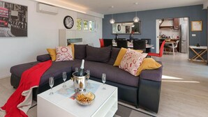 Una sala de estar moderna con un sofá seccional, una mesa de centro con un mapa y aperitivos, una zona de comedor con un espejo, y una cocina visible a través de una puerta abierta.