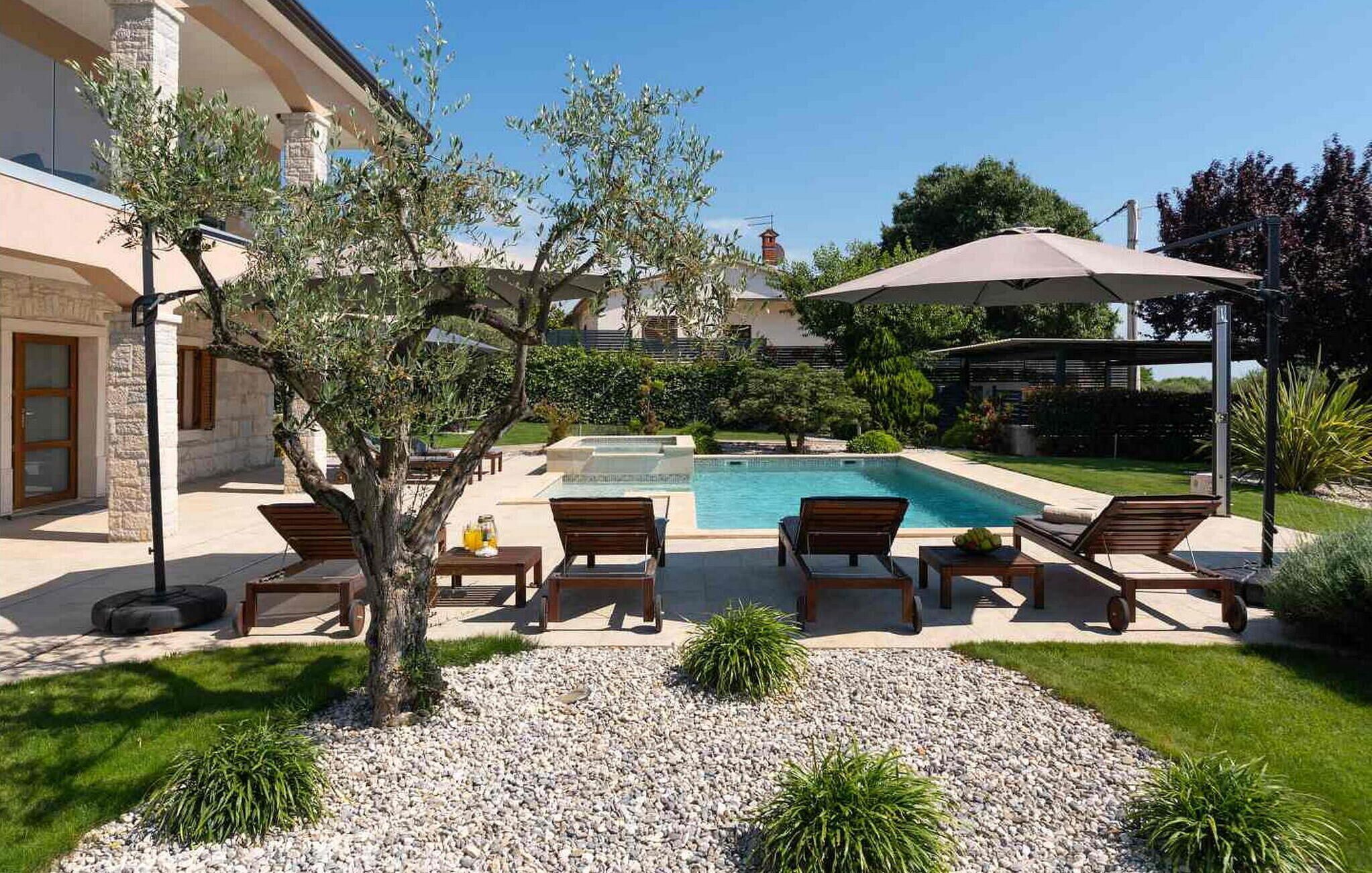 A poolside area with lounge chairs, a pergola with an umbrella, and a stone building in the background.