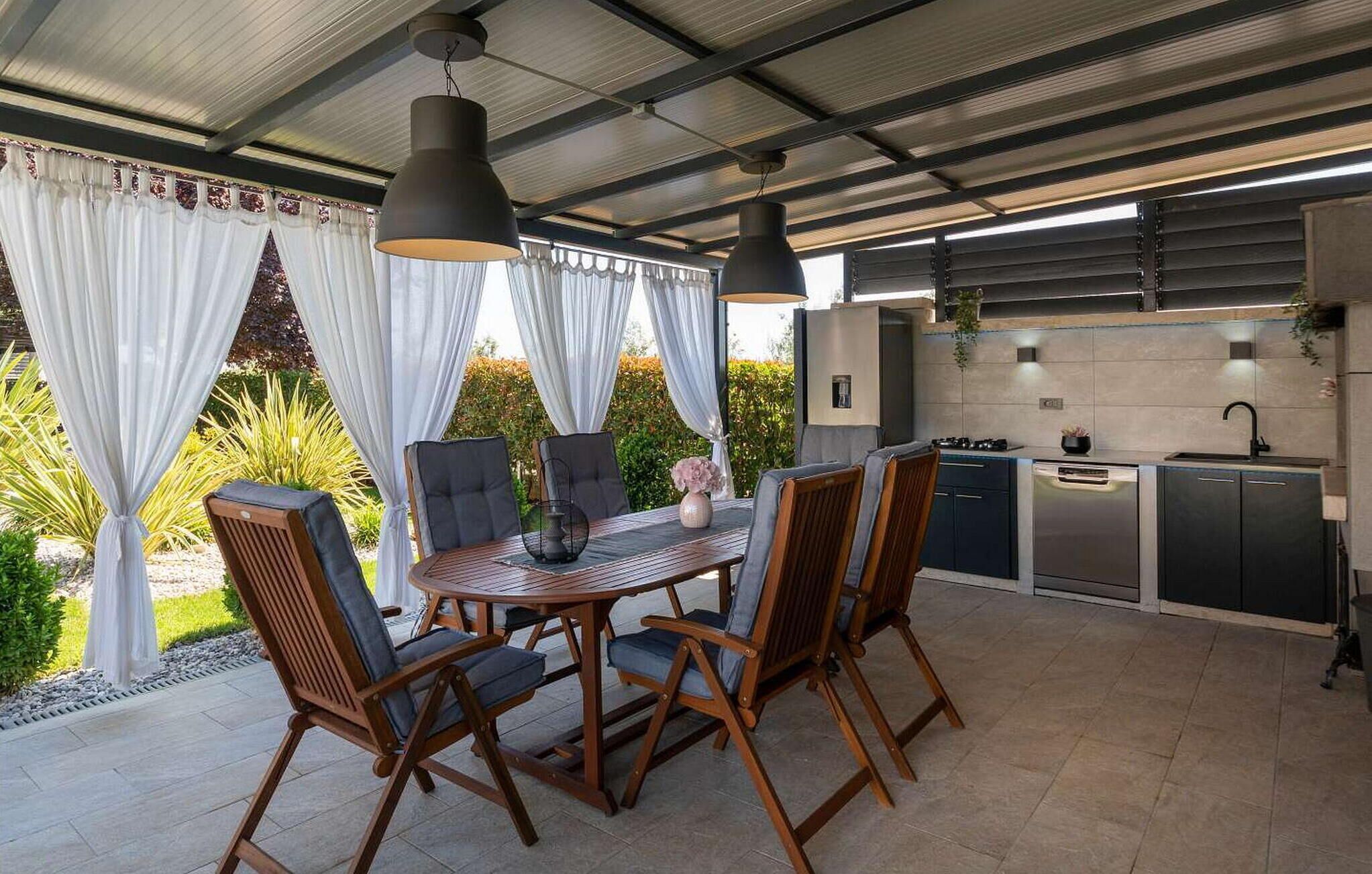 Un patio al aire libre con una mesa redonda y sillas, un colgante de luz y una zona de cocina con fregadero y estufa.