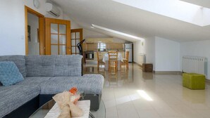 Een ruime woonkamer met een grijze bank, een eettafel met stoelen, een keuken op de achtergrond en een helder, open ontwerp.