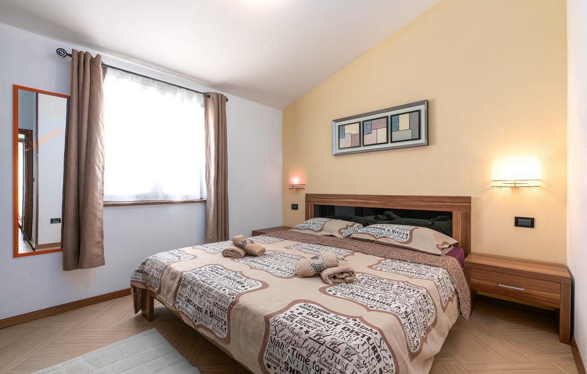 A bedroom with a large bed, a bedside table, a nightstand, a mirror, a window with curtains, and a wall-mounted light fixture.