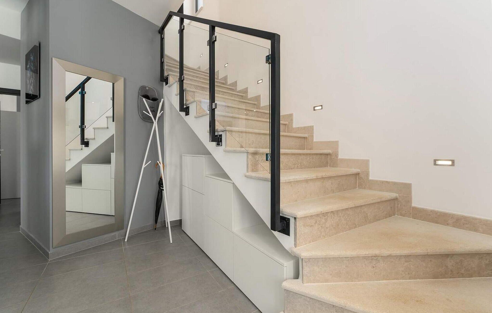 Una escalera moderna con escalones color beige y barandilla de vidrio, que conduce a un rellano con perchero y puerta.
