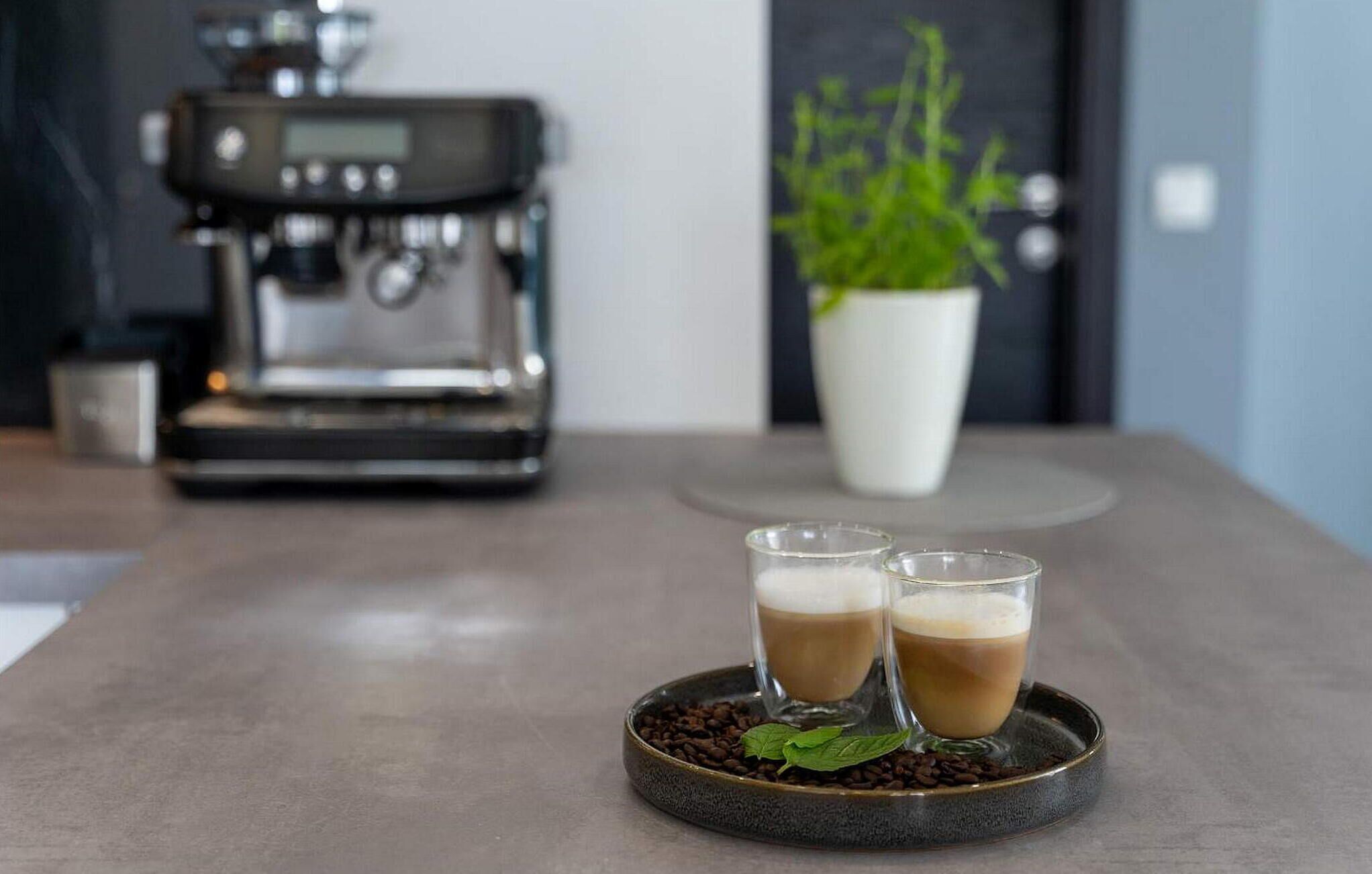 Una cocina moderna con una cafetera y dos tazas de café en una bandeja junto con granos de café y una hoja de menta.