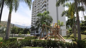Un'area giochi con uno scivolo e strutture per arrampicarsi, circondata da palme e un edificio sullo sfondo.