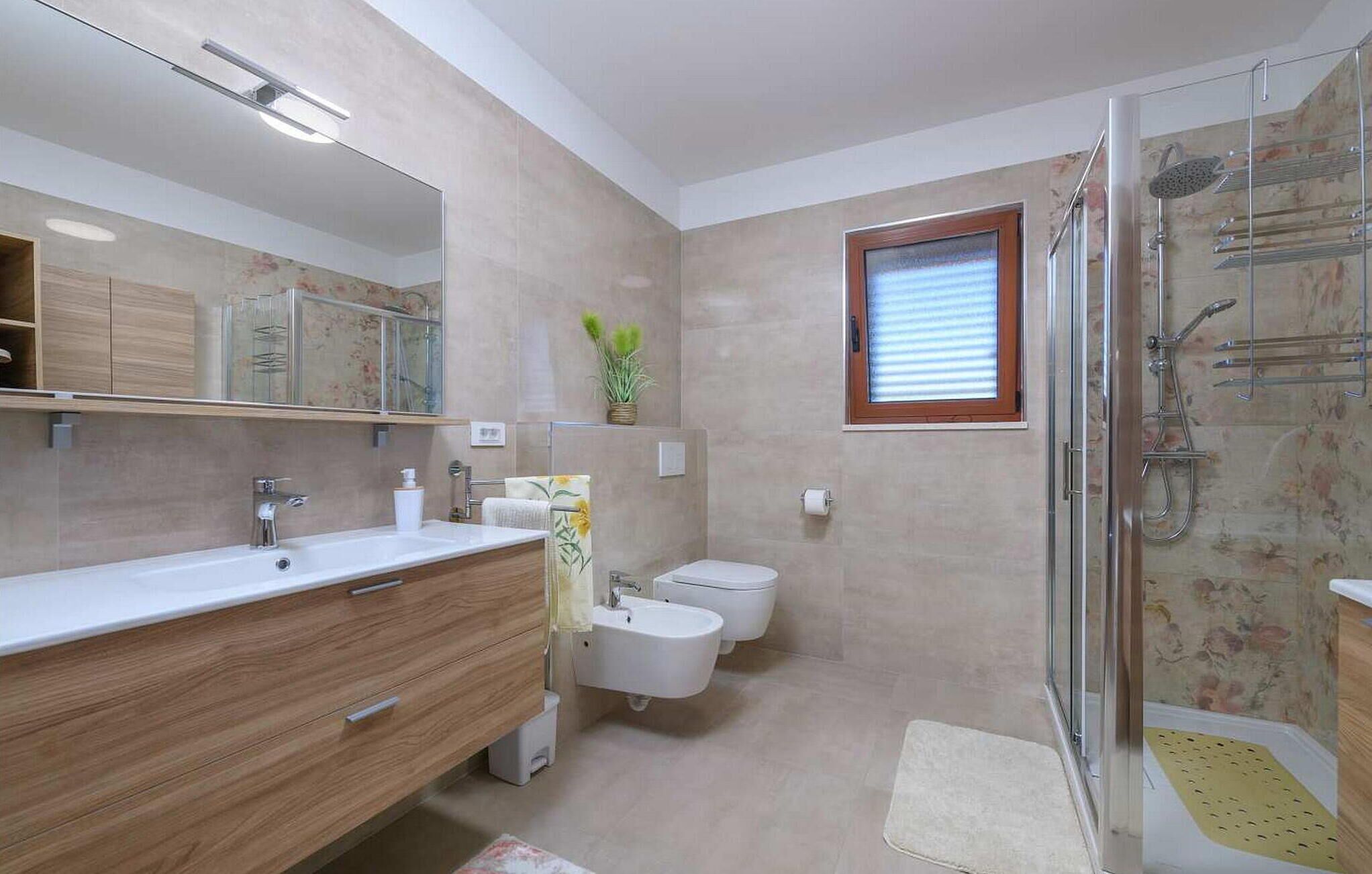 Un baño moderno con un espejo grande, tocador de madera, dos lavamanos, ducha y una ventana pequeña.