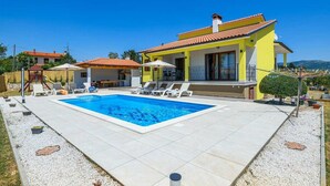 Una villa amarilla de dos pisos con piscina, tumbonas y un porche cubierto.