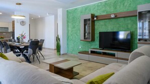 Una sala moderna con una pared verde decorativa, un televisor de pantalla plana, un comedor con mesa y sillas, y una cocina al fondo.