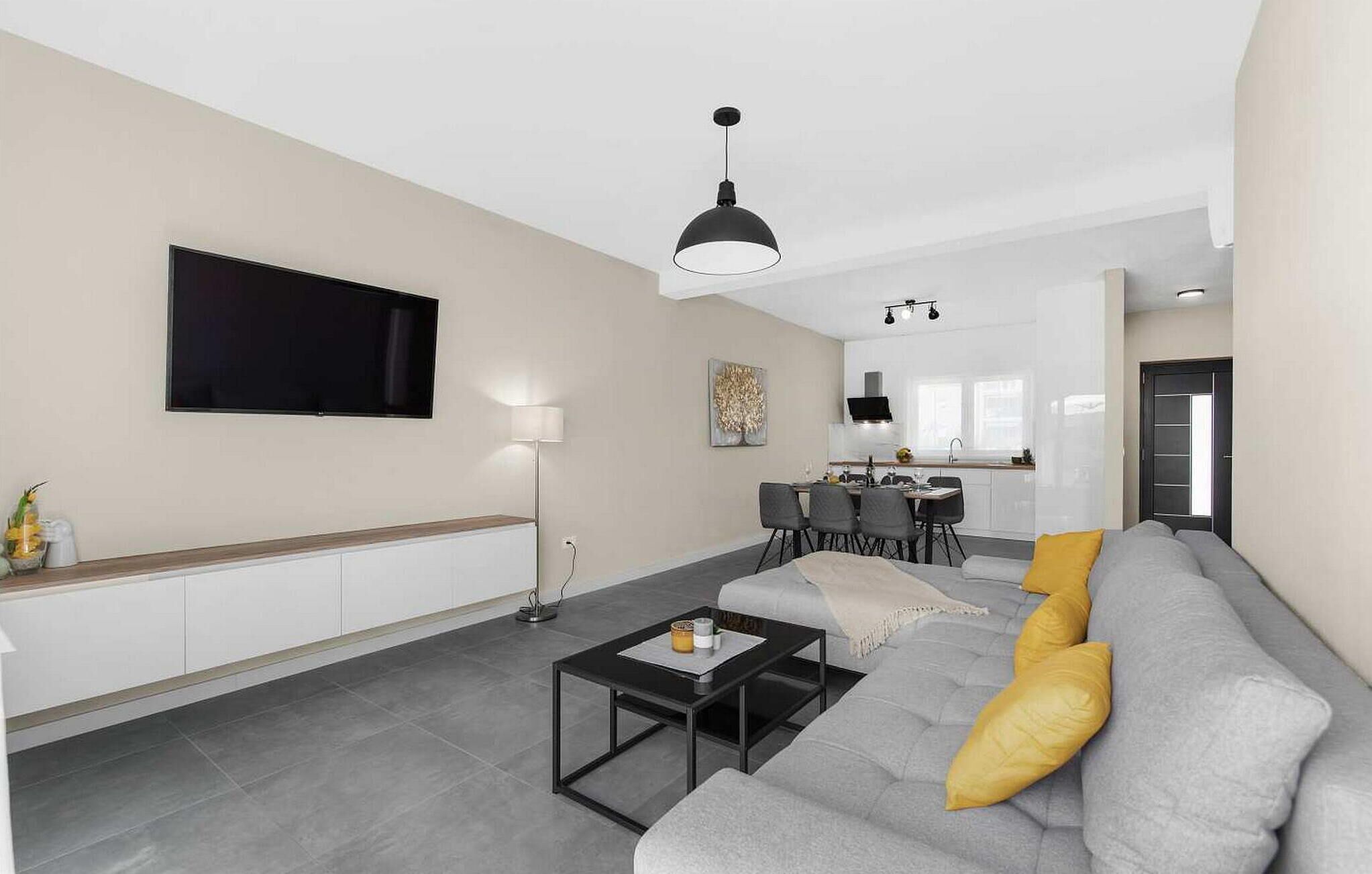 A modern living room with a grey sofa, a coffee table, a flat-screen TV, and a dining area in the background.