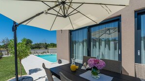 A patio with a table, chairs, and an umbrella. There is a pool in the background. - Awesome home in Porec (Porec)
