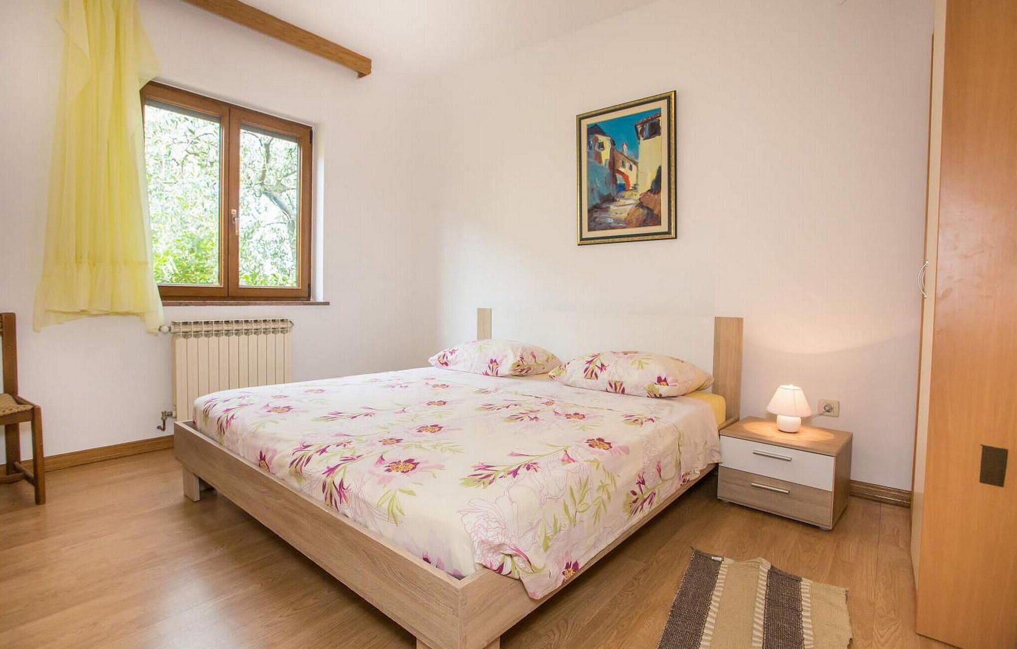Une chambre avec un lit en bois, une table de chevet, une lampe, un radiateur, une fenêtre avec des rideaux et un tableau au mur.