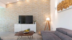 Uma sala de estar moderna com uma parede de pedra, uma TV de tela plana, uma mesa de centro com frutas, um abajur e um sofá cinza.