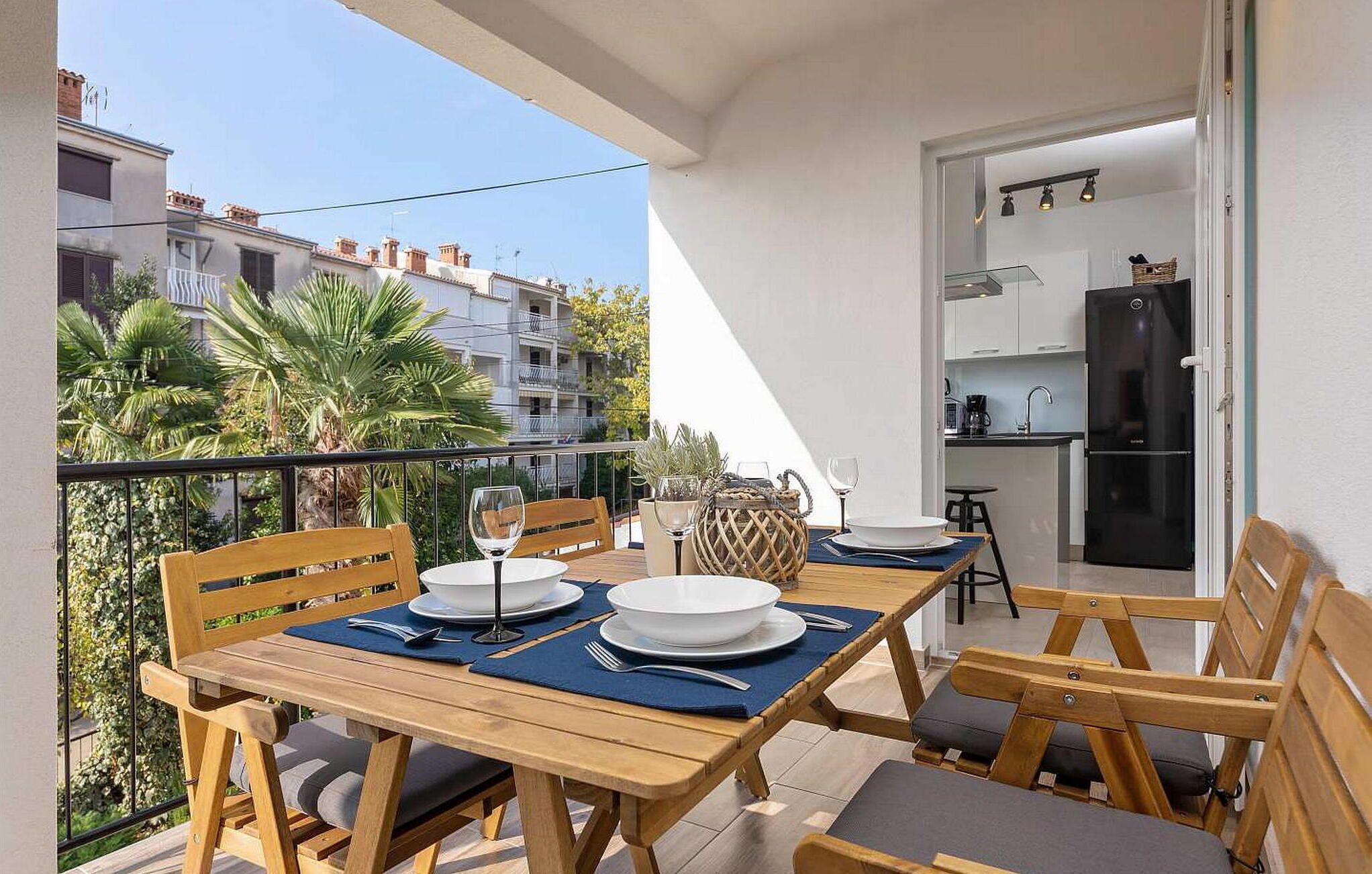 Un balcón con mesa de madera dispuesta para comer, sillas y vistas a edificios y follaje cercanos.