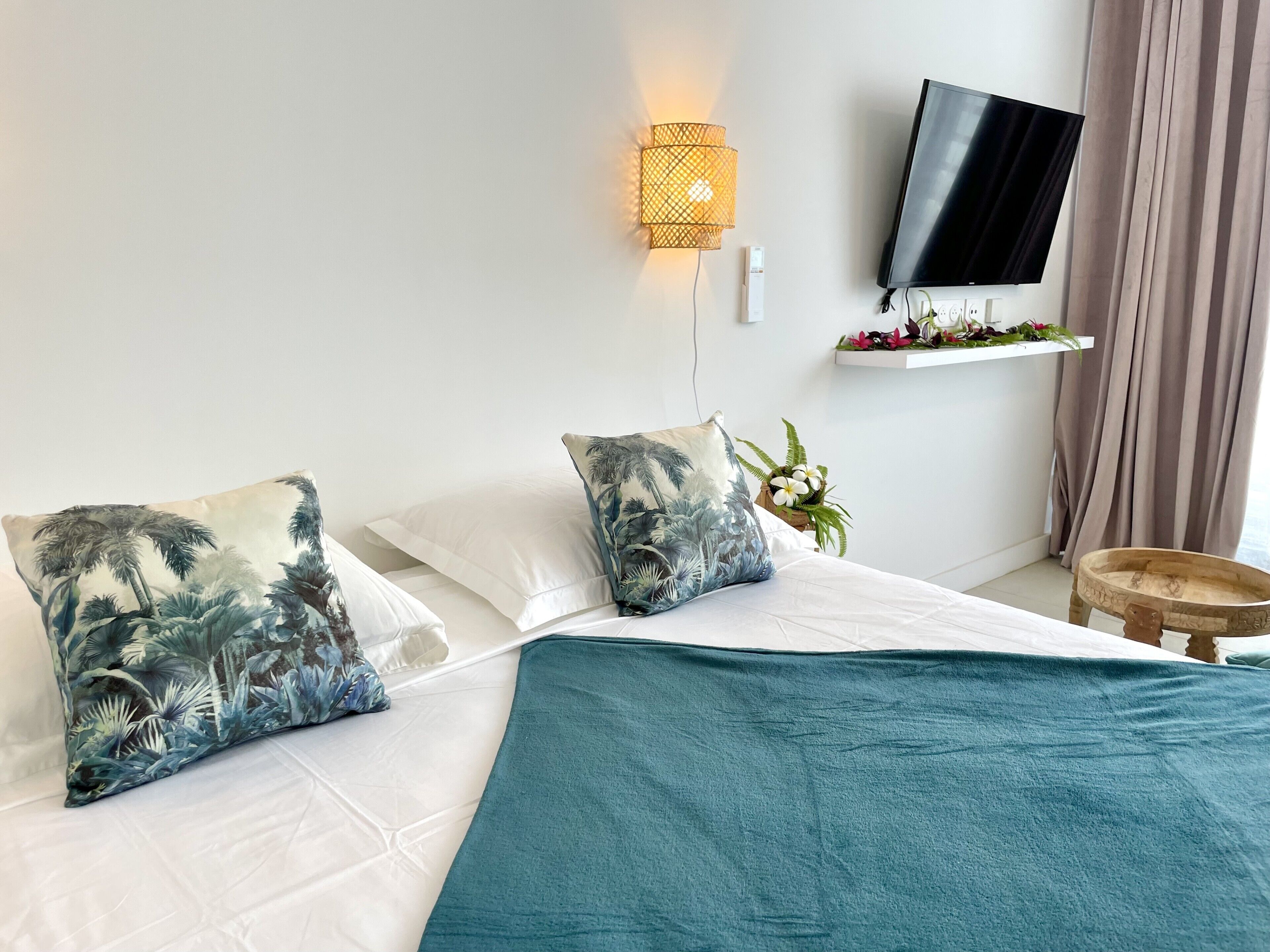 Un lit bien fait, recouvert d’une couverture turquoise, avec deux oreillers à motifs tropicaux, un téléviseur fixé au mur et une petite étagère ornée de plantes.