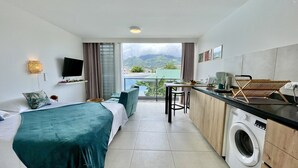 Habitación de hotel con cama, una kitchenette pequeña, lavadora y balcón con vistas a las montañas.