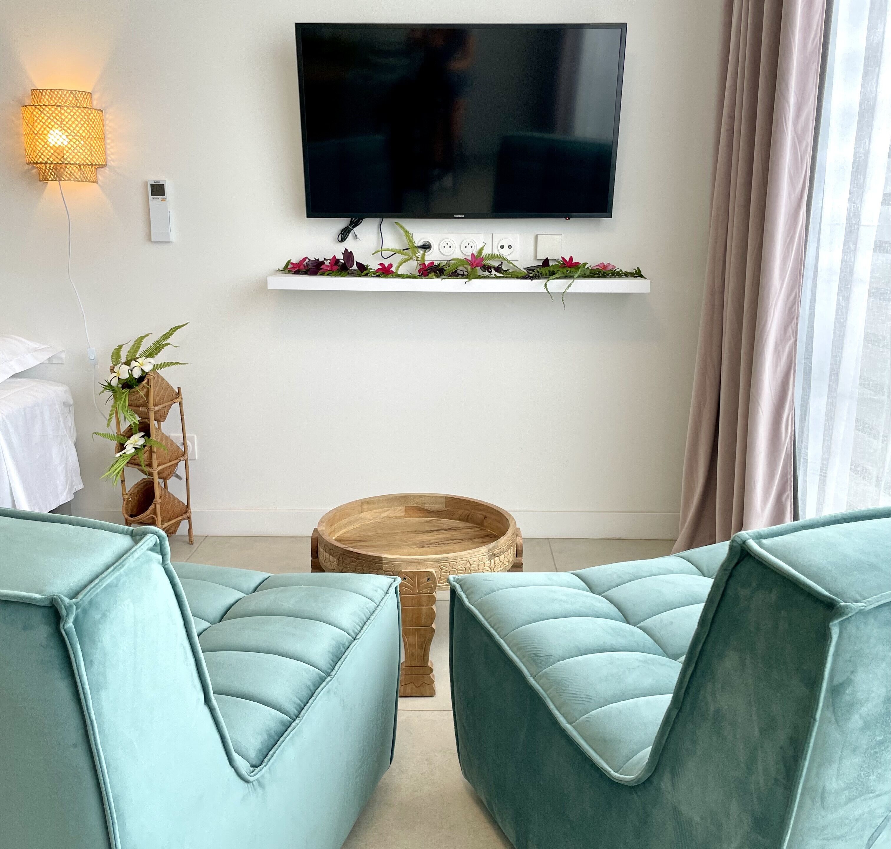 Un salon moderne avec des canapés de couleur turquoise, un téléviseur fixé au mur, une étagère décorative agrémentée de fleurs, une table d'appoint en osier et une table basse ronde en bois.