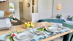 Habitación de hotel con una mesa de comedor dispuesta con platos, un frutero y un vaso. Se observa una cama con una colcha verde y almohadas, y una lámpara fijada a la pared.