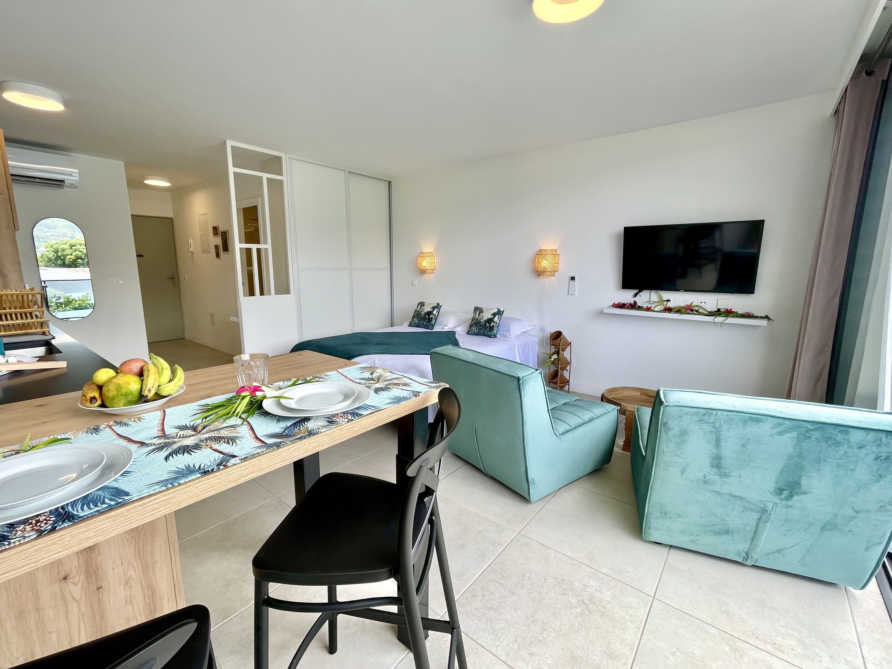 Un salon moderne avec un canapé, une table à manger ornée de fruits et d’un verre, et un téléviseur fixé au mur.