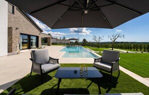 Ein moderner Poolbereich im Freien mit zwei Stühlen und einem Tisch, umgeben von einem gepflegten Rasen und einem Steinhaus.