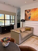 Una sala de estar con un sofá color beige, un mueble negro, un cuadro grande y un florero con flores.