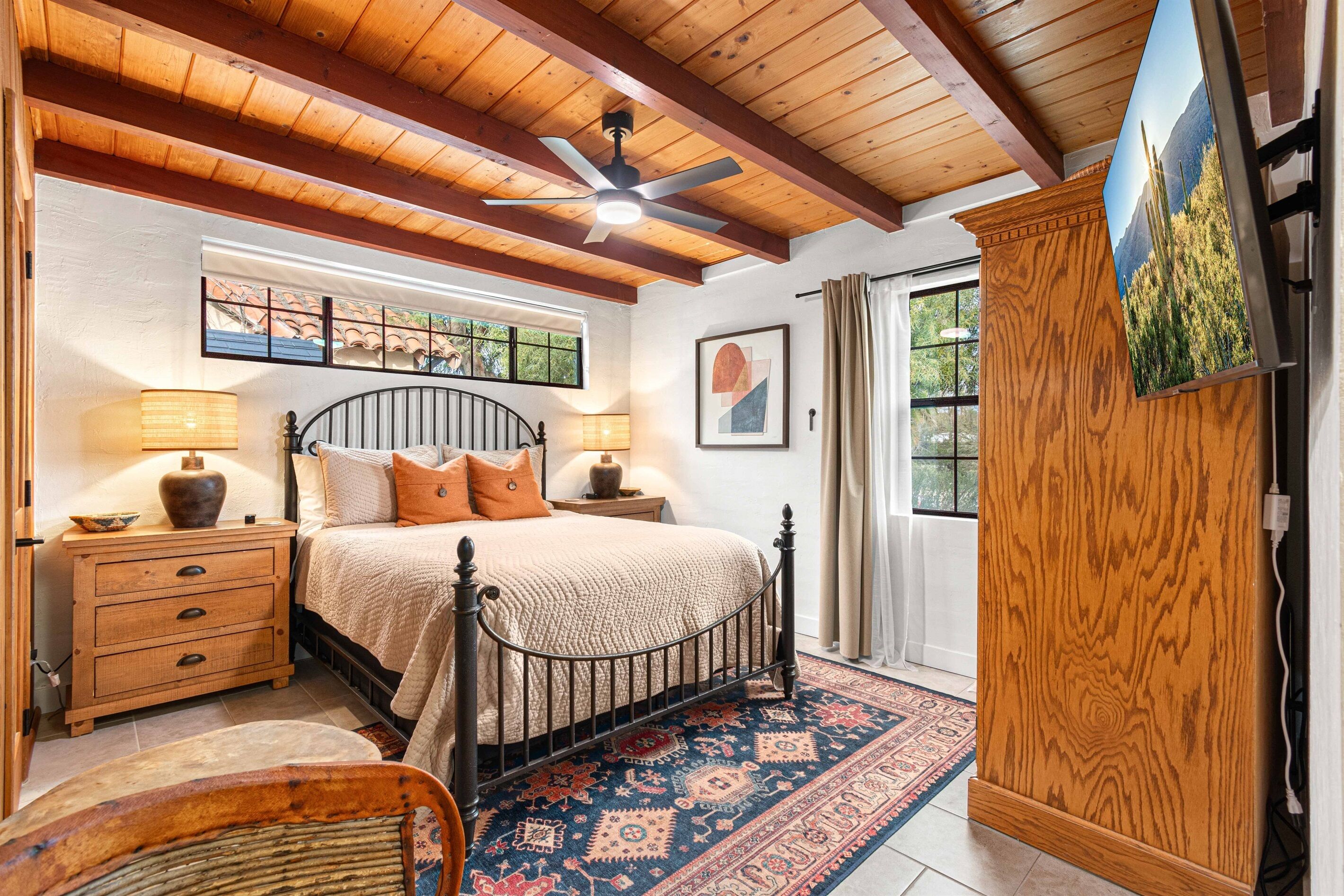 A bedroom with a bed, wooden headboard, a ceiling fan, a nightstand with a lamp, a dresser, and a window with curtains.